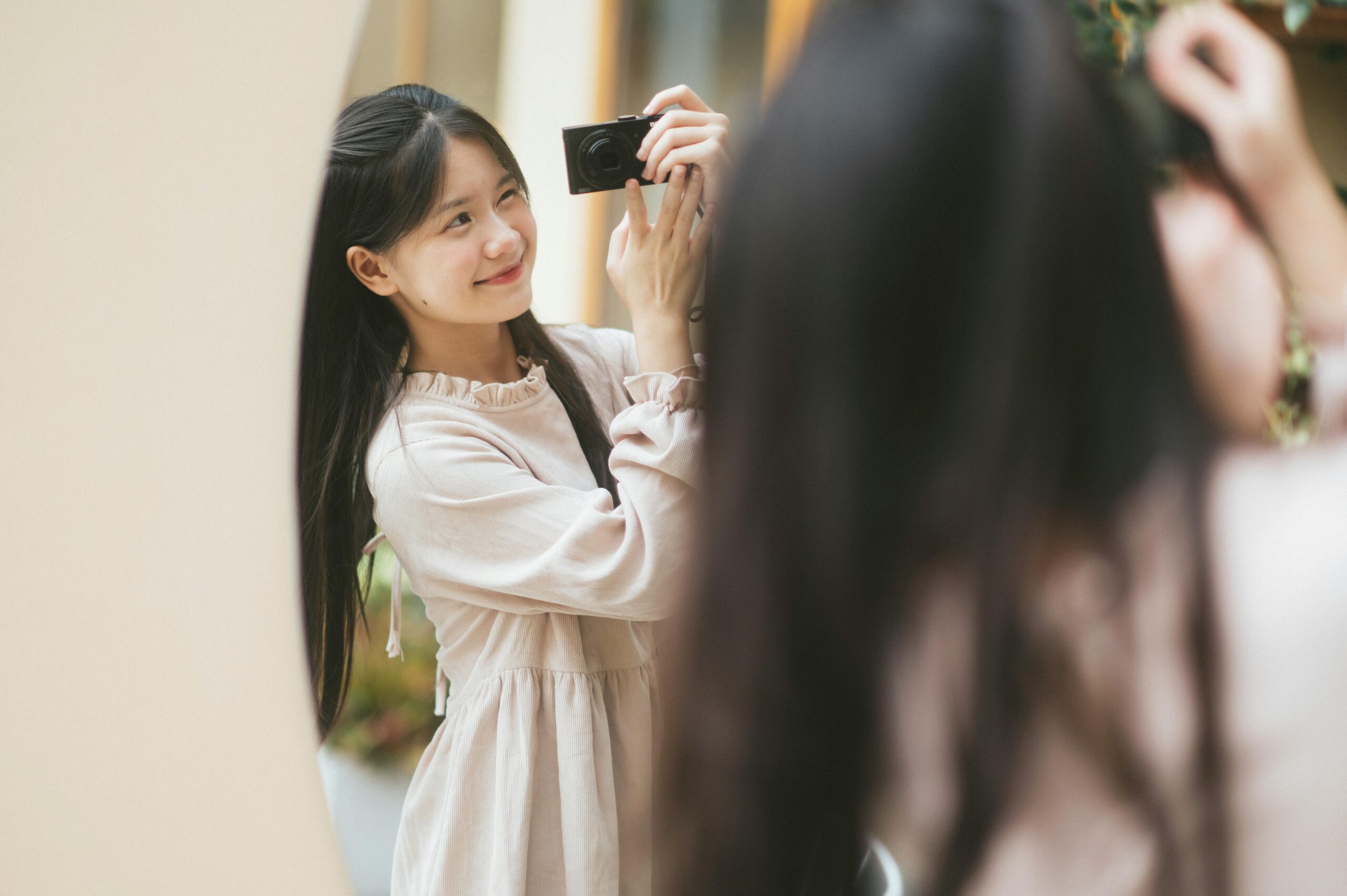 A young woman in a dress smiles while taking a selfie with a camera in front of a mirror.